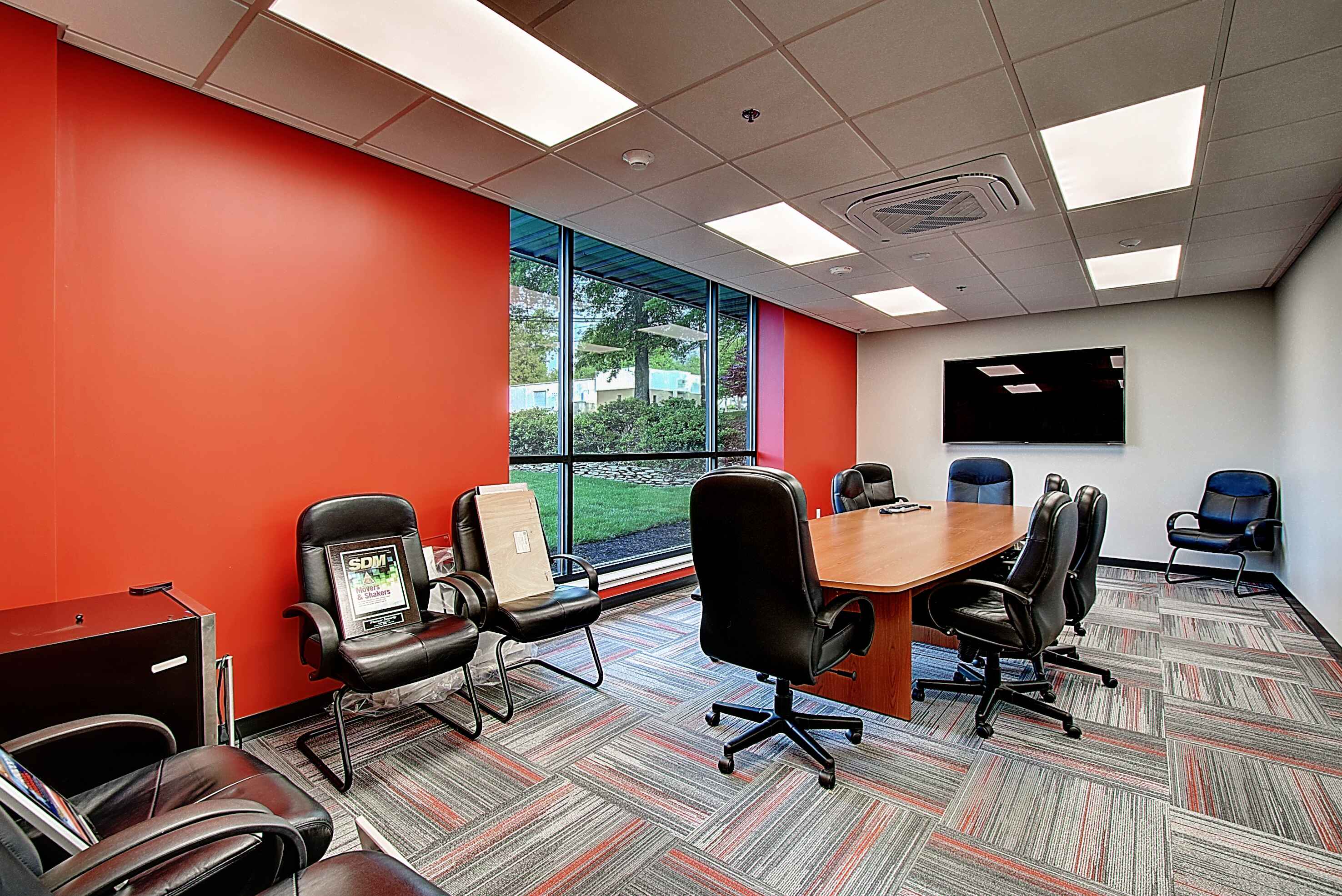 Modern meeting room featuring a large wooden table, black chairs, orange walls, and a window with natural light.