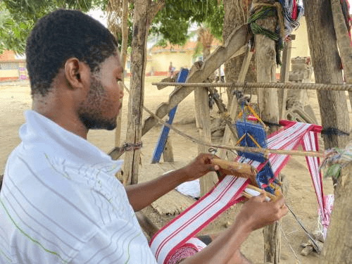 Element Craftsmanship of traditional woven textile Kente (Ghana) Inscribed in 2024 (19.COM) on the Representative List of the Intangible Cultural Heritage of Humanity Title EN: Weaver weaving a strip of Kente. Here he passes the loaded weft shuttle through the partition made by the hurdles and he pulls his legs upwards and downwards. With the beater, he beats the woven yarns for a firm hold.