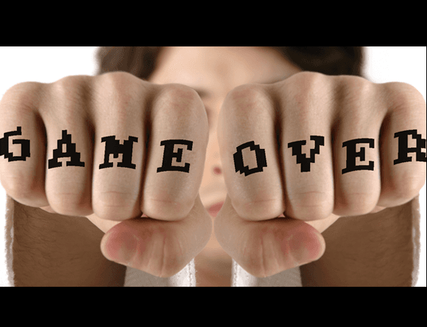 The image shows two hands clasped together, with fingers extended and forming a fist shape. Each finger is marked with the word "GAME" in black capital letters, arranged in a straight line across the palm. The hands are positioned in front of a blurred face, with no other features visible. The background is uniformly white.