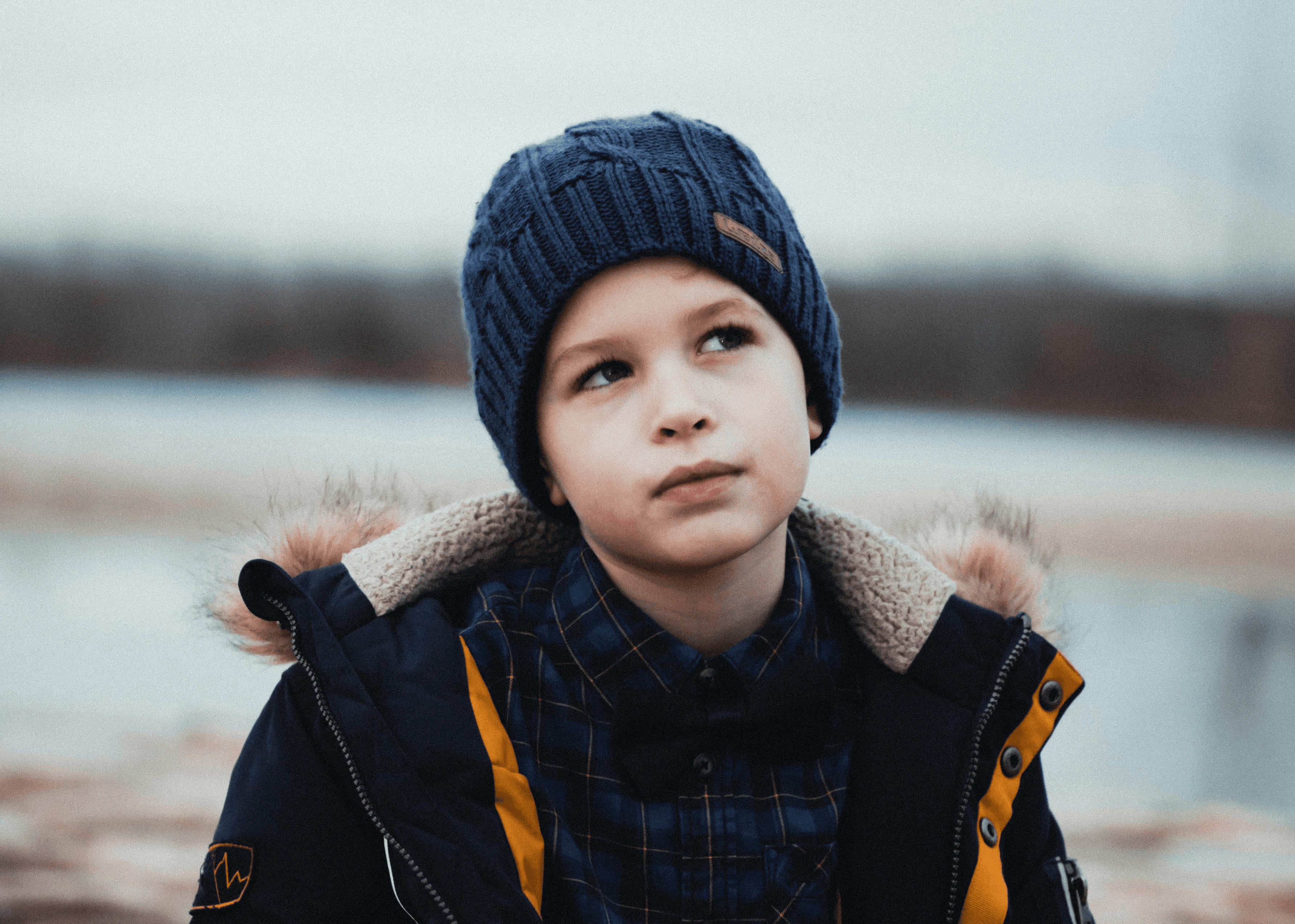 boy in black and yellow jacket and knit cap standing near body of water during daytime