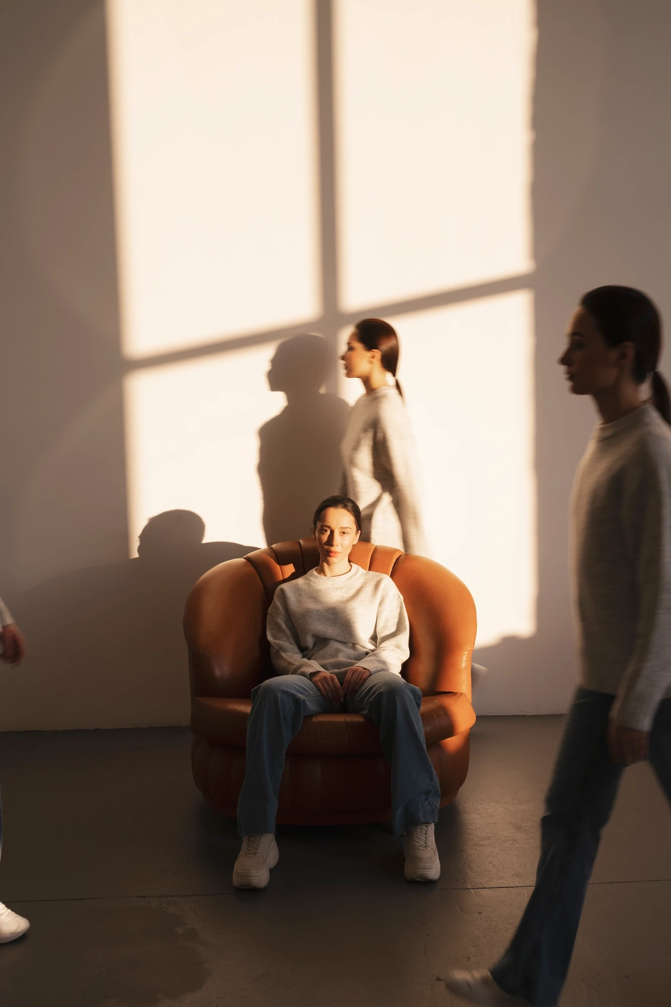 A woman sitting on a chair in a room with sunlight on the wall, and another woman walking around her
