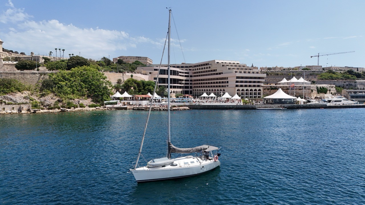 A Beneteau First 38.5 sailing yacht at anchor near a waterfront hotel and beach club in St. Julian's, Malta, with its mast raised and a boom cover fitted.