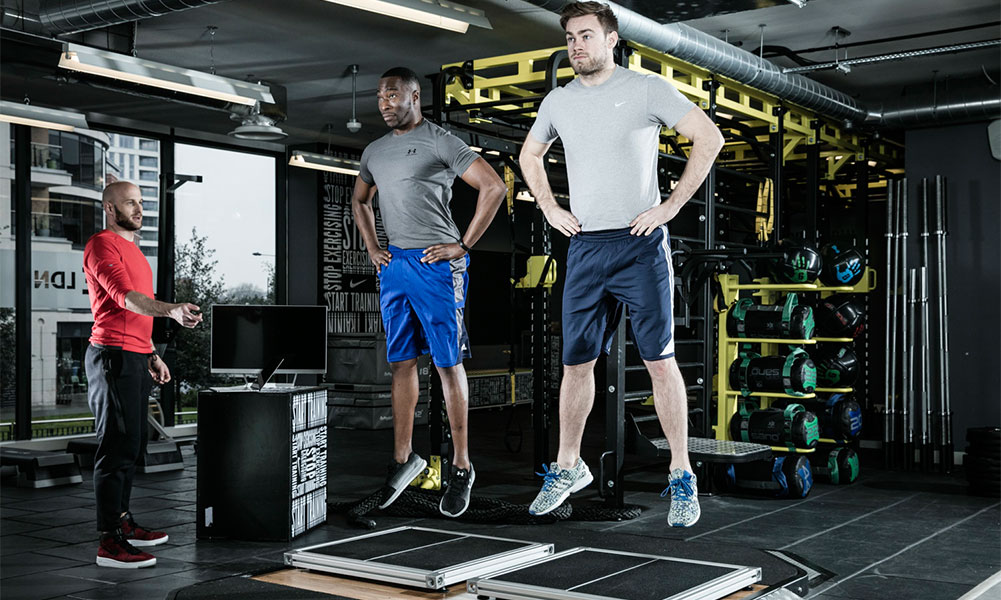 Two male athletes performing vertical jump testing under coach supervision in a modern training facility—focused on explosive power and performance tracking.