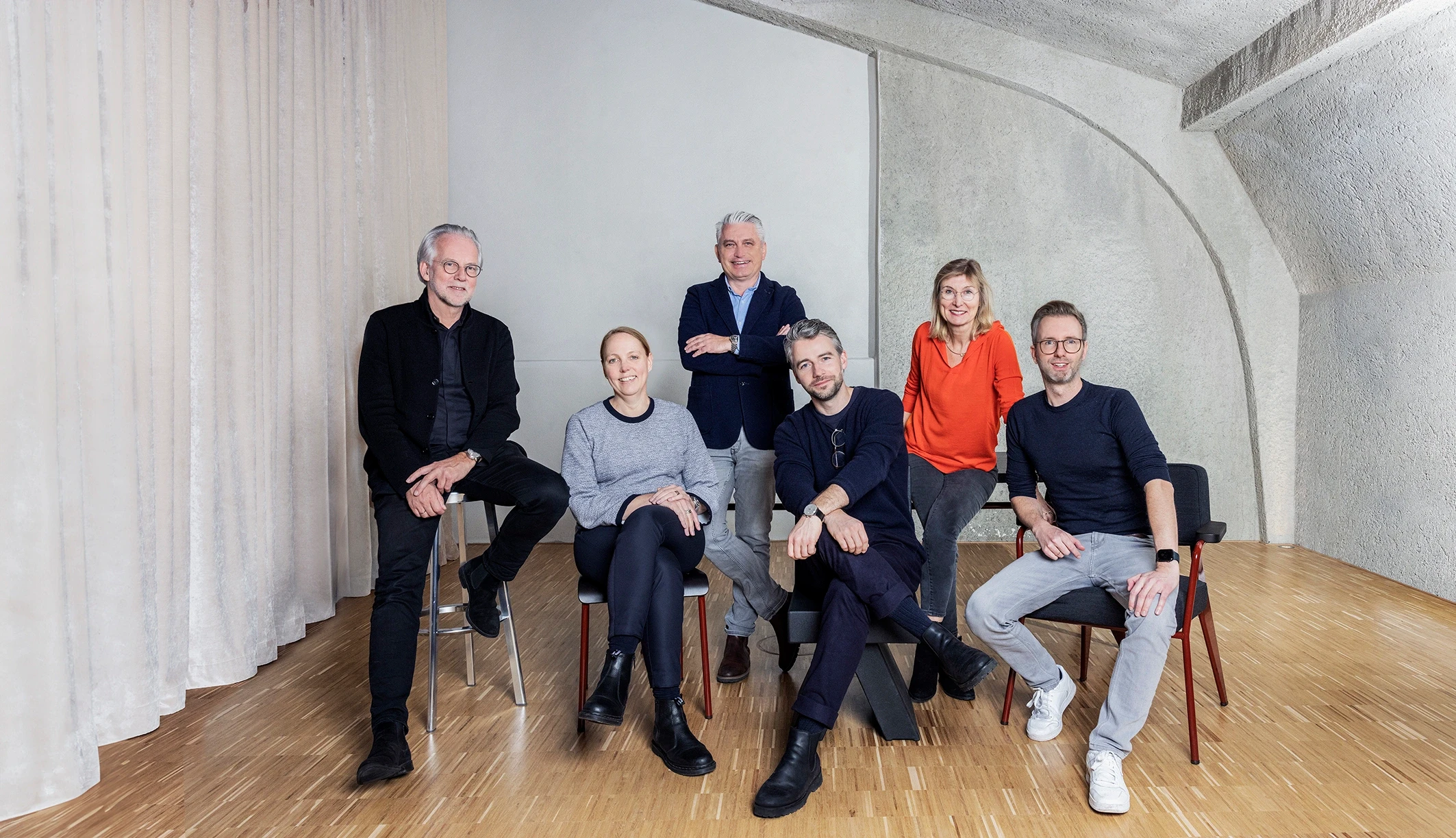 Gruppenfoto des MAAS & PARTNER Teams in einem modernen Innenraum mit Sichtbetondecke, Vorhängen und Holzfußboden.