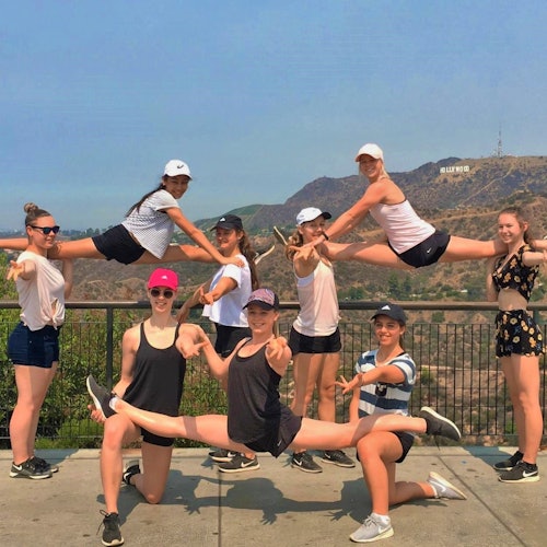 Group of nine people posing energetically near a fence with the Hollywood sign in the background. Two are mid-air in splits.