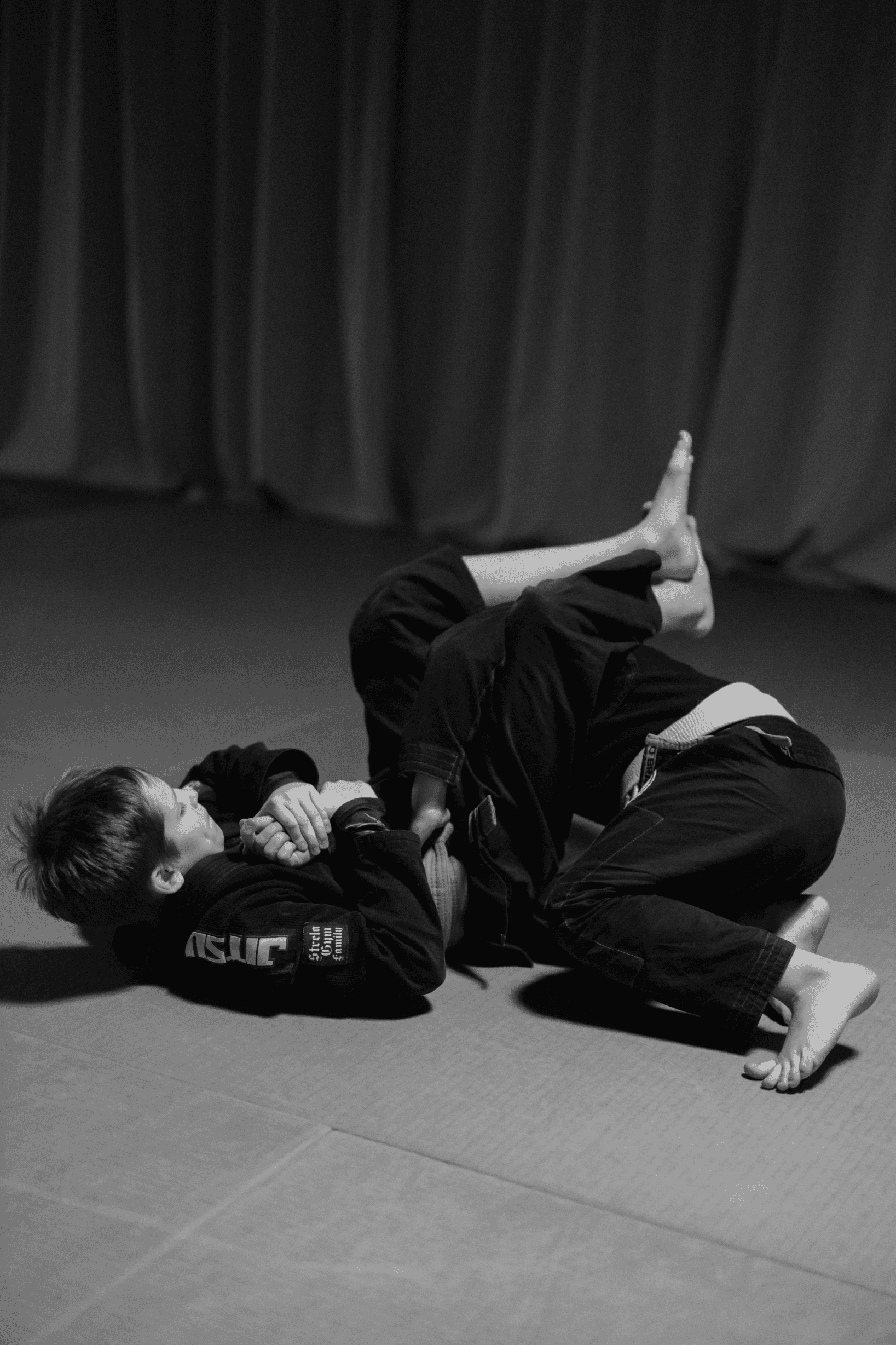 Two children wearing black gis practicing Jiu-Jitsu ground techniques on a grey mat, with one student's yellow belt visible during the grapple.
