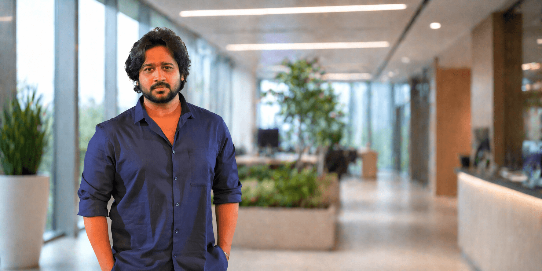 Man in navy shirt standing in modern office space with plants and blurred corporate background.