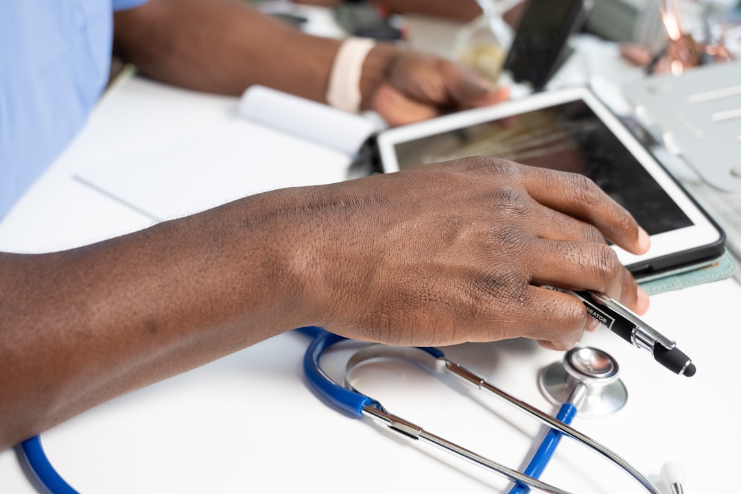 Close-up of a clinician's hands typing on a laptop with a high-tech AI dashboard visible on a secondary monitor, stethoscope sitting on the desk, soft natural light
