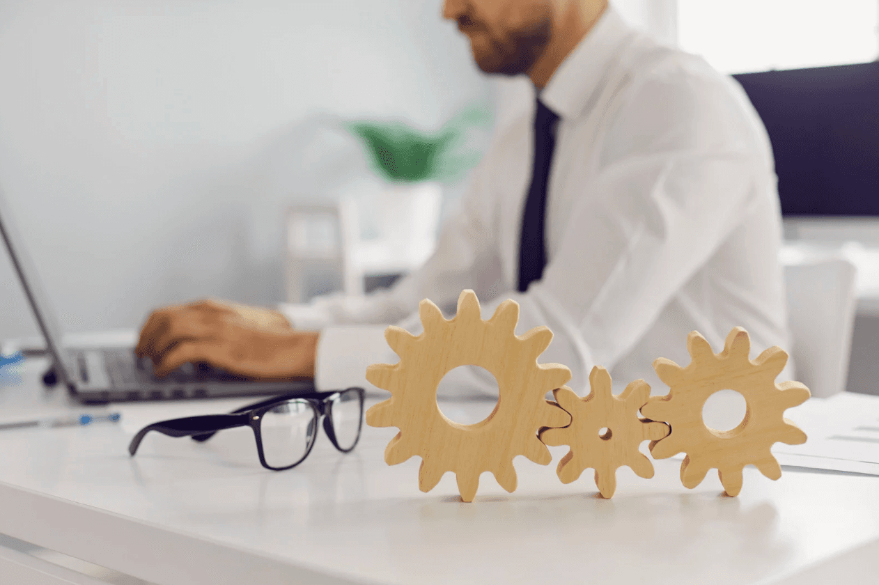 Business professional working at a laptop with wooden gear cogs on the desk — representing ASIN-level automation and finely tuned Amazon campaign performance.