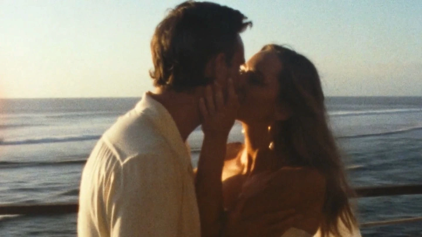 A couple sharing a romantic kiss on a beach at sunset, with the ocean waves gently rolling in the background under a clear sky.