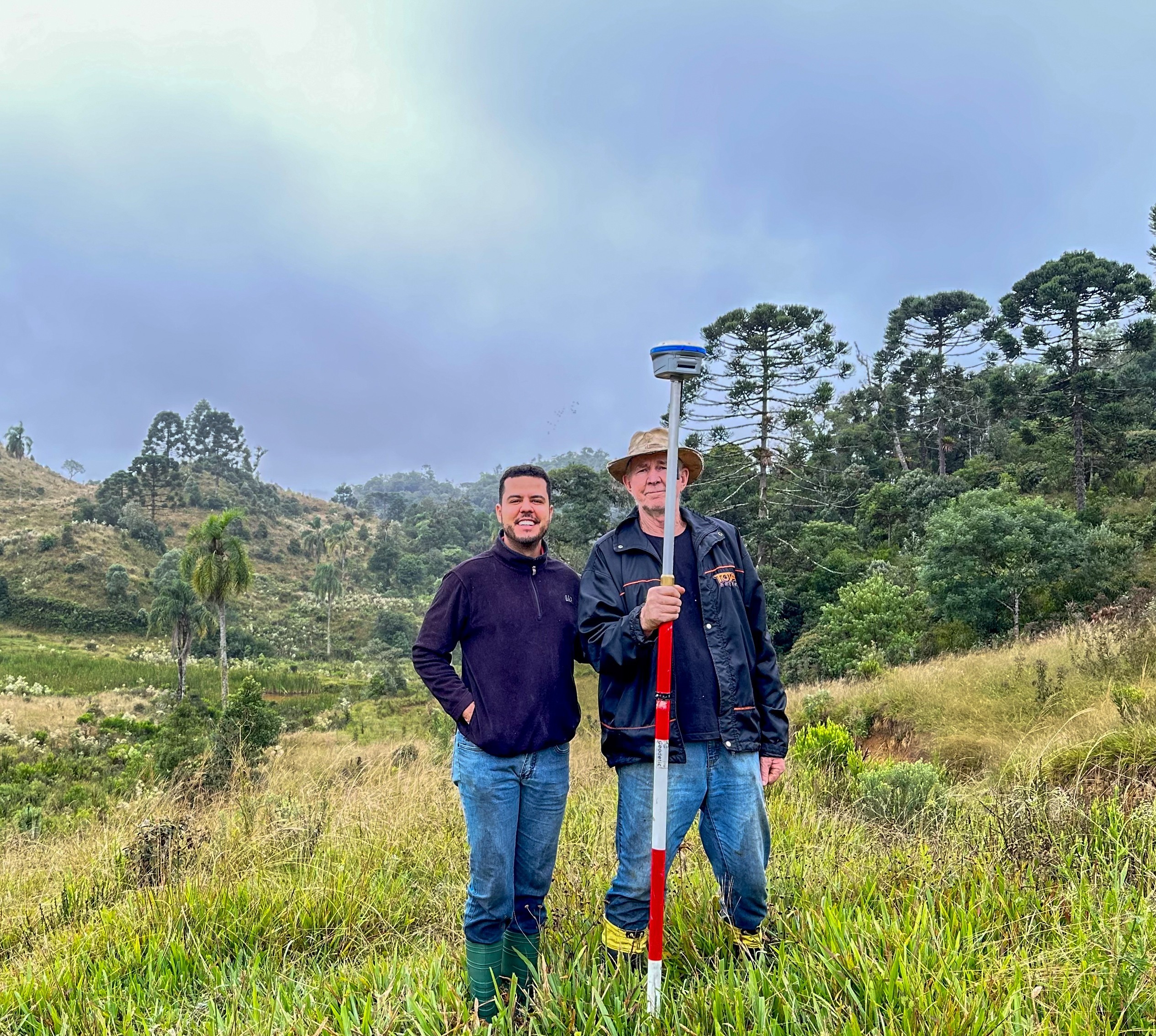 Dois profissionais de engenharia em campo aberto com relevo montanhoso, segurando um receptor GNSS RTK para levantamento topográfico de precisão.