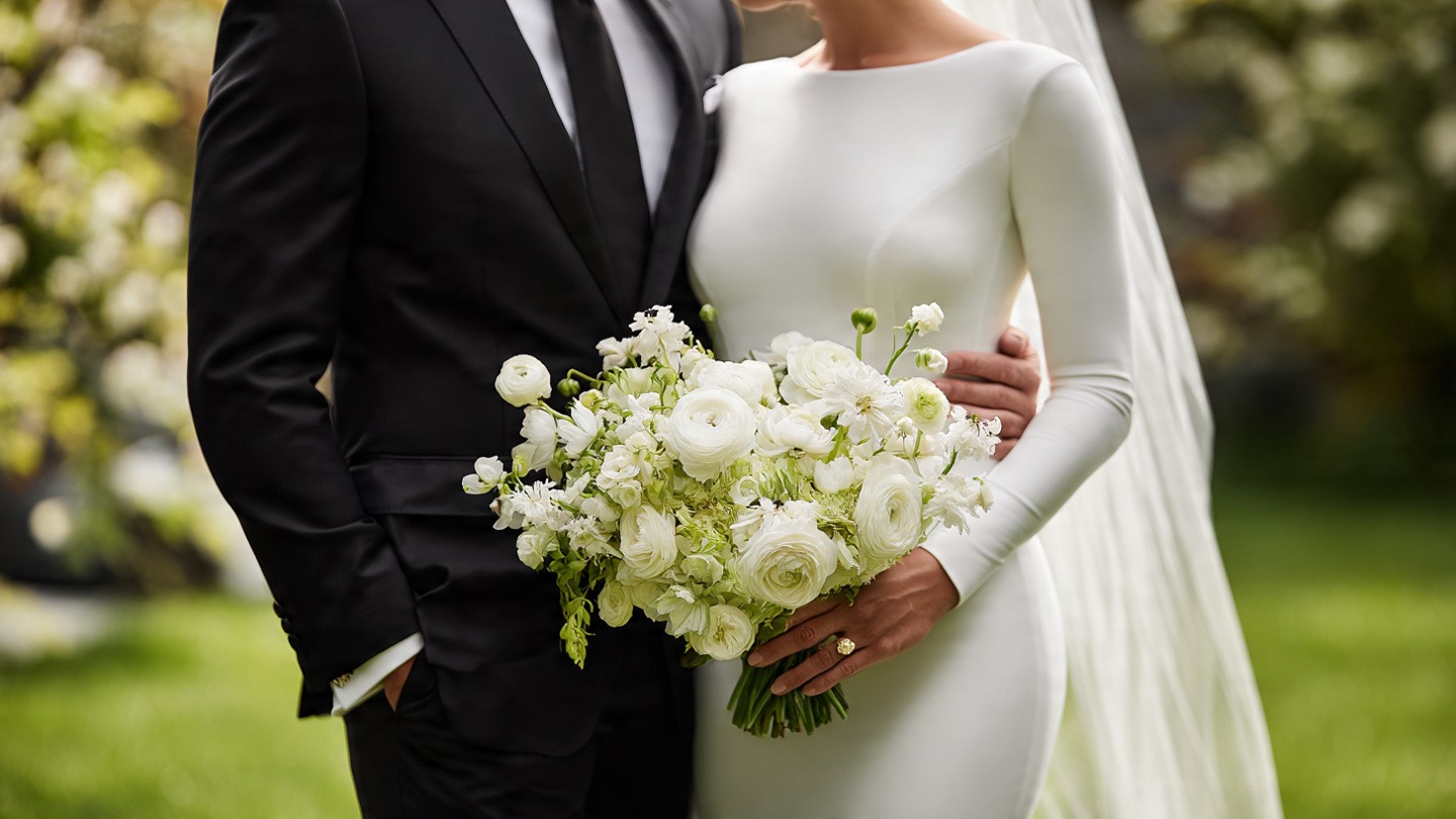 Intimate portrait of a bride and groom on grass; the groom in a black tuxedo with bow tie gently kisses the bride’s forehead as she holds a generous bouquet of white ranunculus, poppies, and foliage against her form-fitting long-sleeve ivory wedding dress and flowing veil.