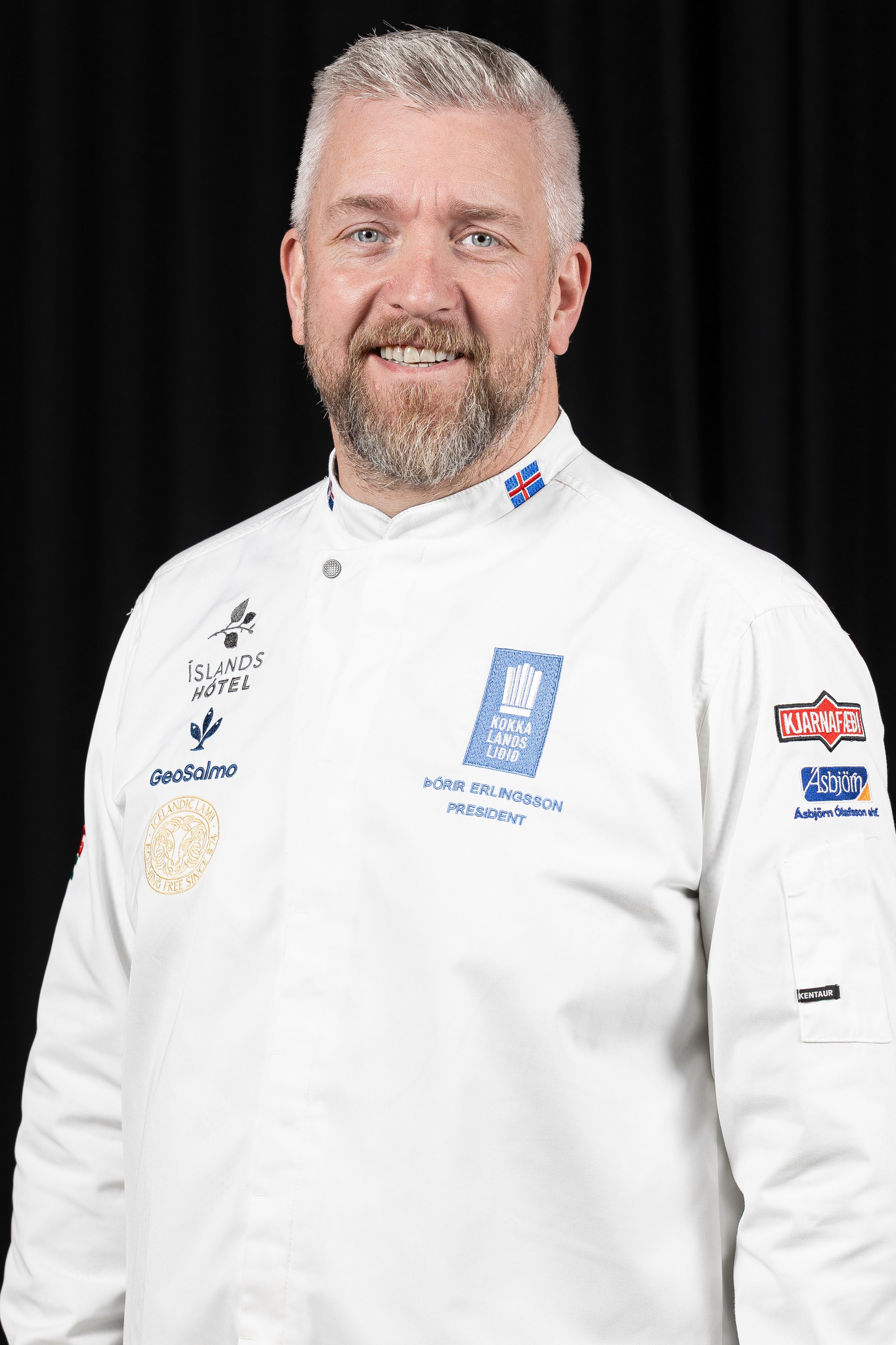 Thorir Erlingsson smiles in a portrait, wearing the white jacket of the Icelandic National Culinary Team embroidered with his name and the title President, against a black background.
