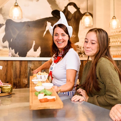 一位女士和一个女孩在柜台前，桌上有木板盛放的食物，背景是牛的壁画，头顶上方悬挂着吊灯。
