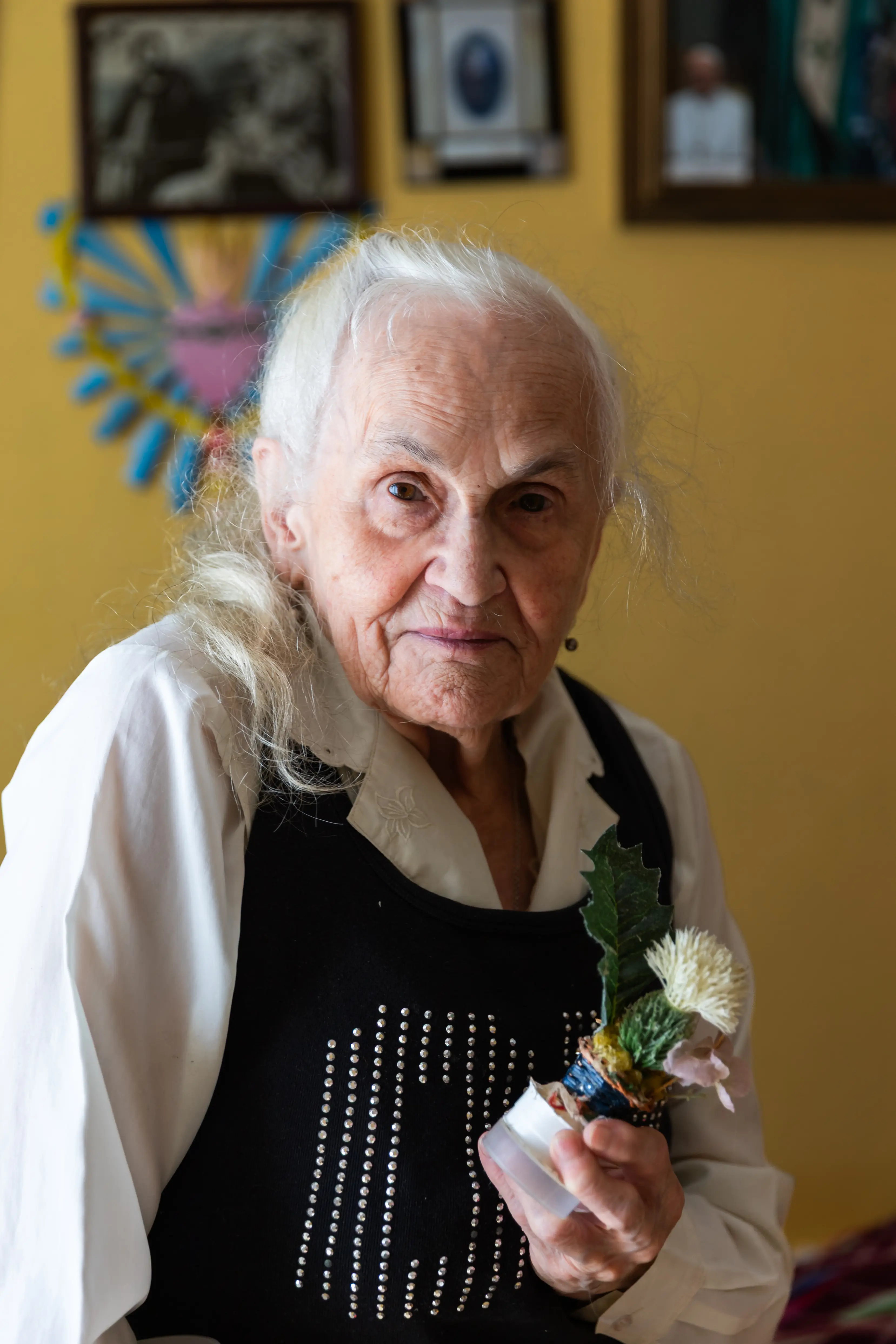 Starsza kobieta o długich siwych włosach trzyma w ręku kwiatek. Jest ubrana w białą koszulę i kamizelkę. W tle ściana z obrazami.