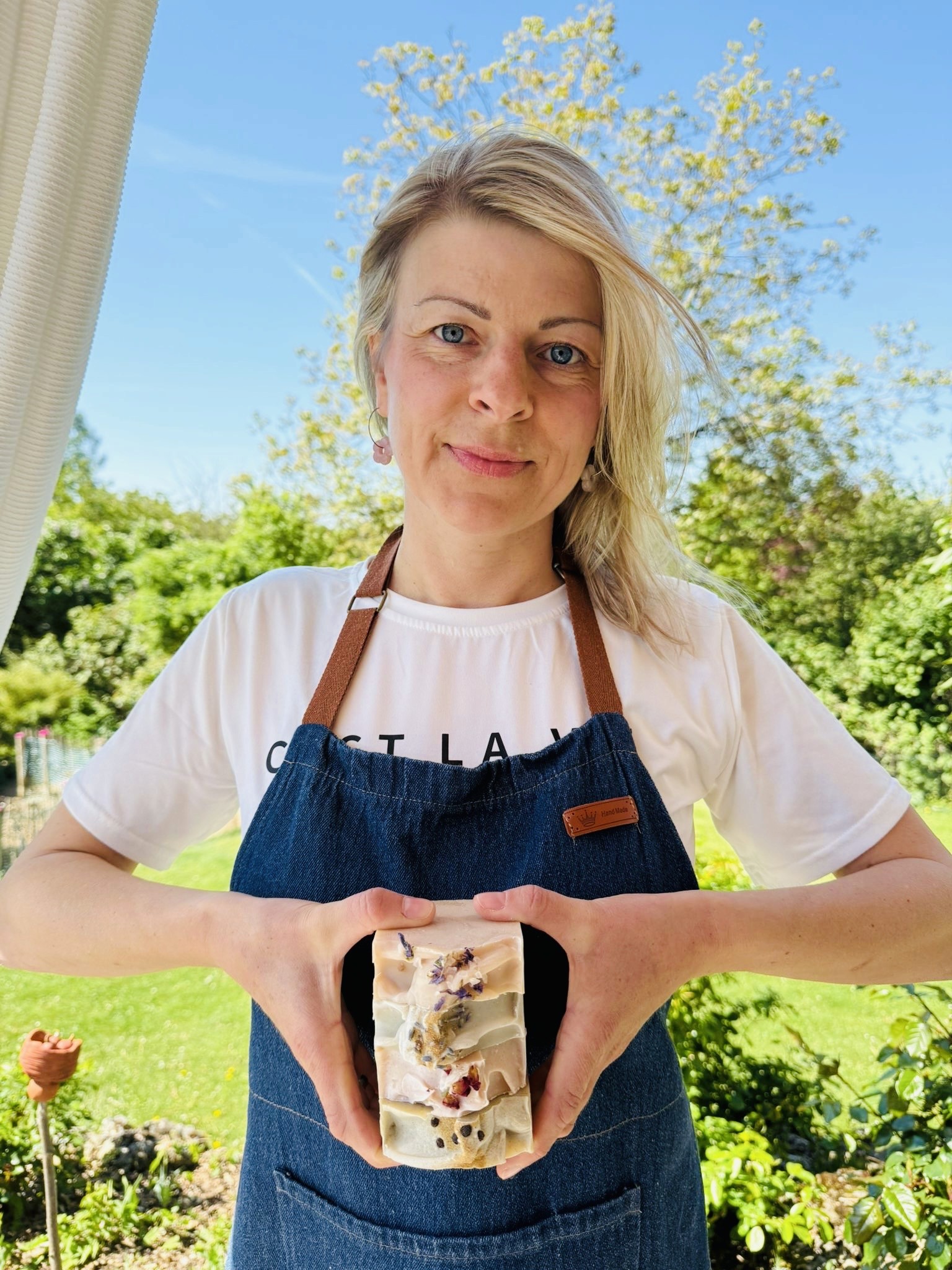 Die Gründerin der Kalous Seifenmanufaktur, Sonja Heumann, hält handgemachte Naturseifen mit Blütenverzierungen in ihren Händen. Fotografiert im Garten bei Sonnenschein.