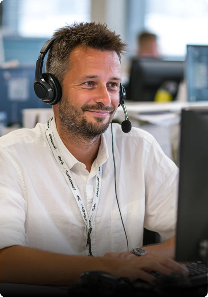 A man with short brown hair and a beard, wearing a white shirt and a black headset with a microphone, smiles while looking at a computer screen in a bright office environment.