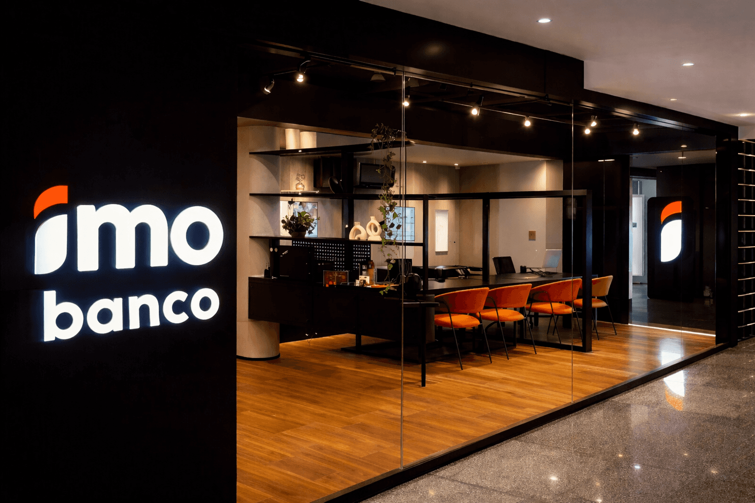 Fotografia da fachada e interior da agência do Imobanco, com letreiro luminoso à esquerda. O espaço é visível através de uma vitrine de vidro e apresenta um design moderno com piso de madeira, quatro cadeiras cor terracota e decoração minimalista com plantas.