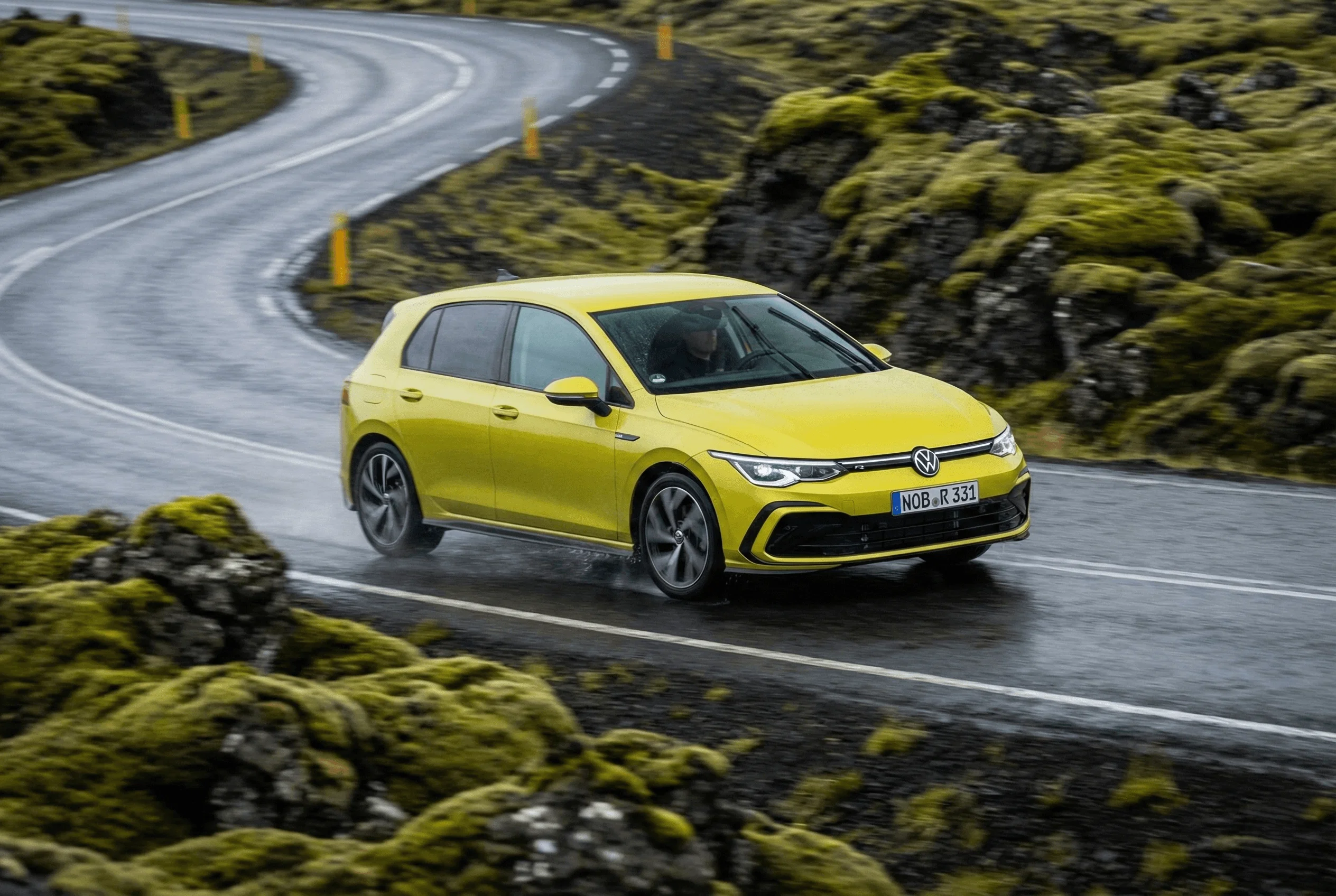 A yellow VW Golf driving on a wet asphalt road winding through a mossy lava field.