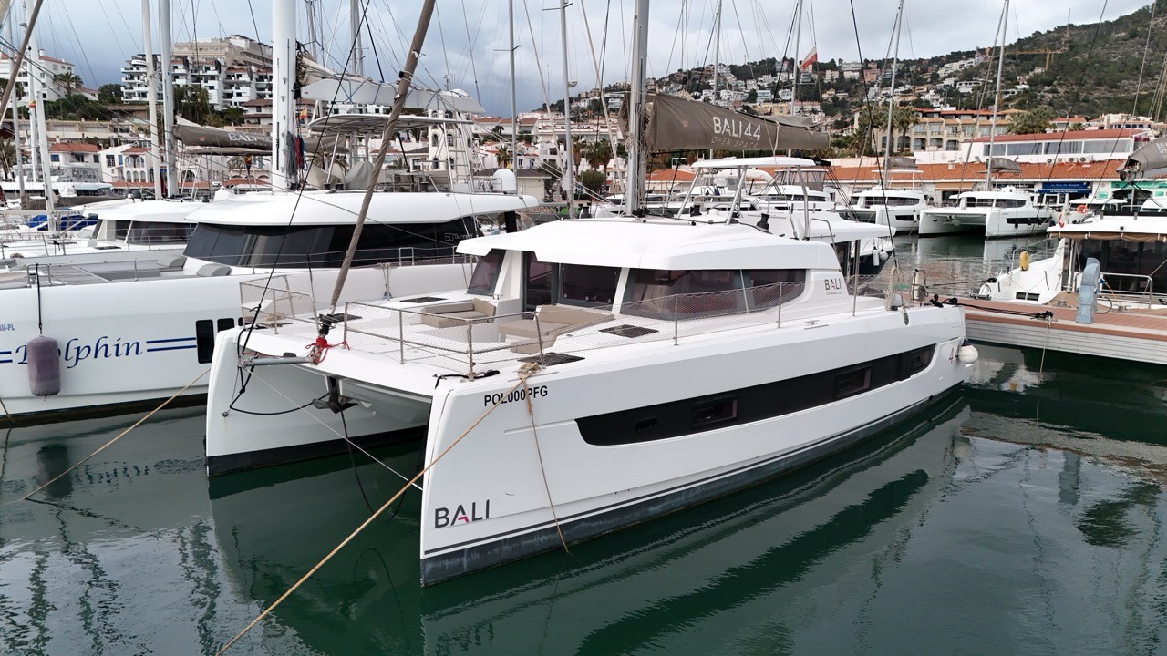 Port bow three-quarter view of a Bali 4.4 sailing catamaran berthed in a busy Mediterranean marina, showing the sleek white twin hulls, panoramic saloon windows, open forward cockpit net, hardtop roof, and Bali branding on the port hull bow, with hillside town visible in the background