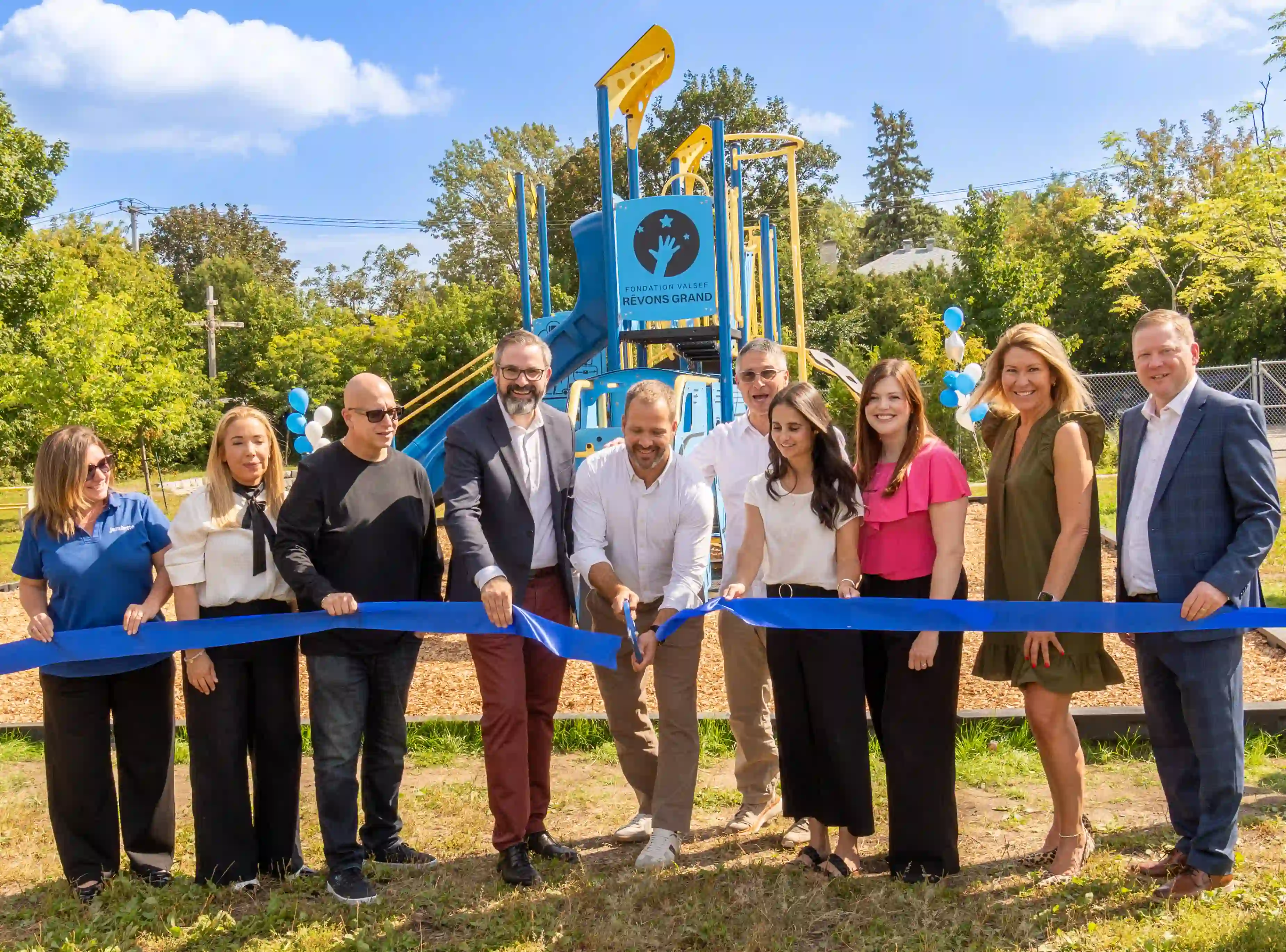 Un groupe de membres de la Valsef Dream Big Foundation et de représentants de l'école coupant un ruban bleu devant un nouveau terrain de jeu bleu et jaune à l'école Cardinal-Léger par une journée ensoleillée