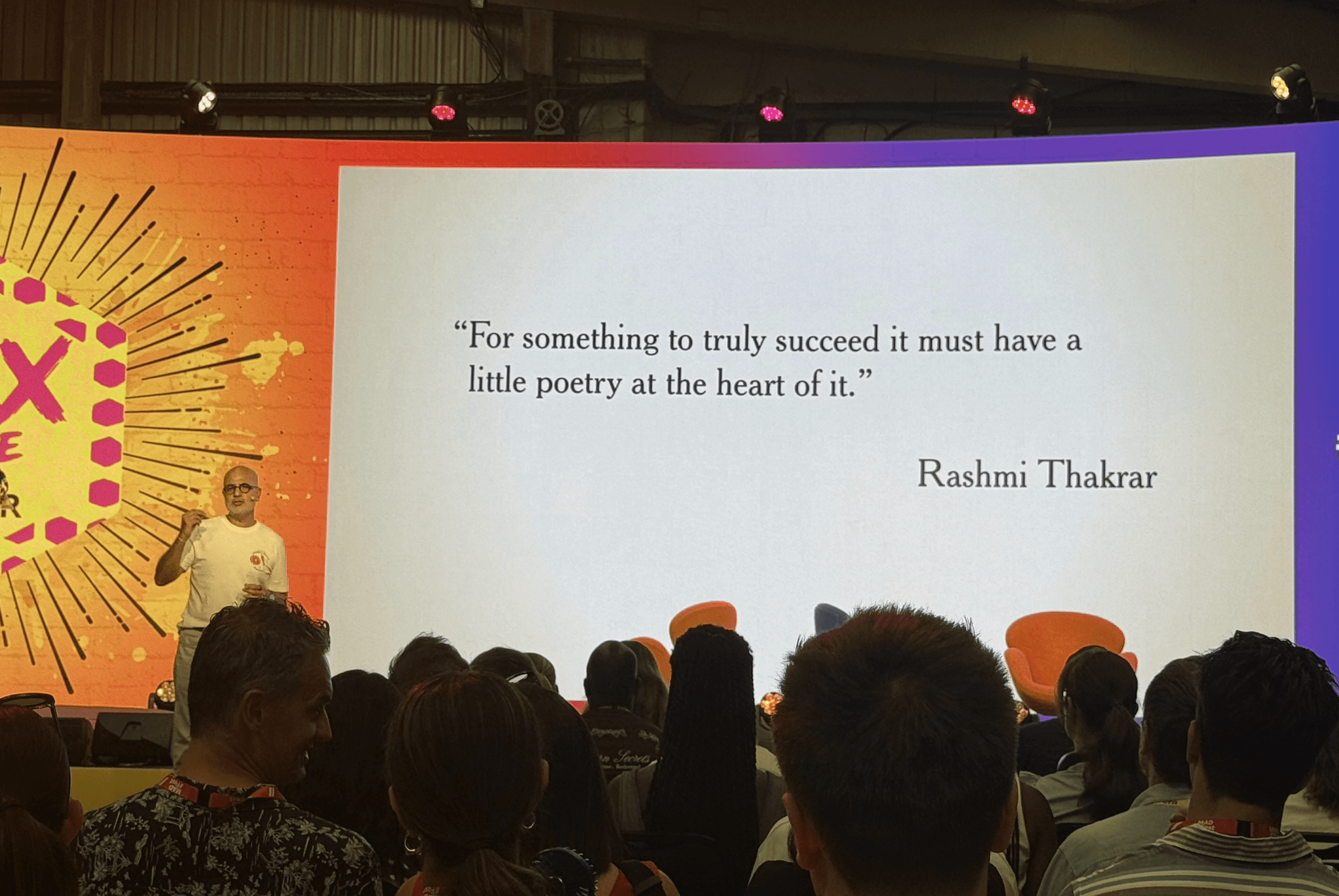 A speaker stands beside a large screen displaying the quote 'For something to truly succeed it must have a little poetry at the heart of it' attributed to Rashmi Thakrar, in front of a seated audience.