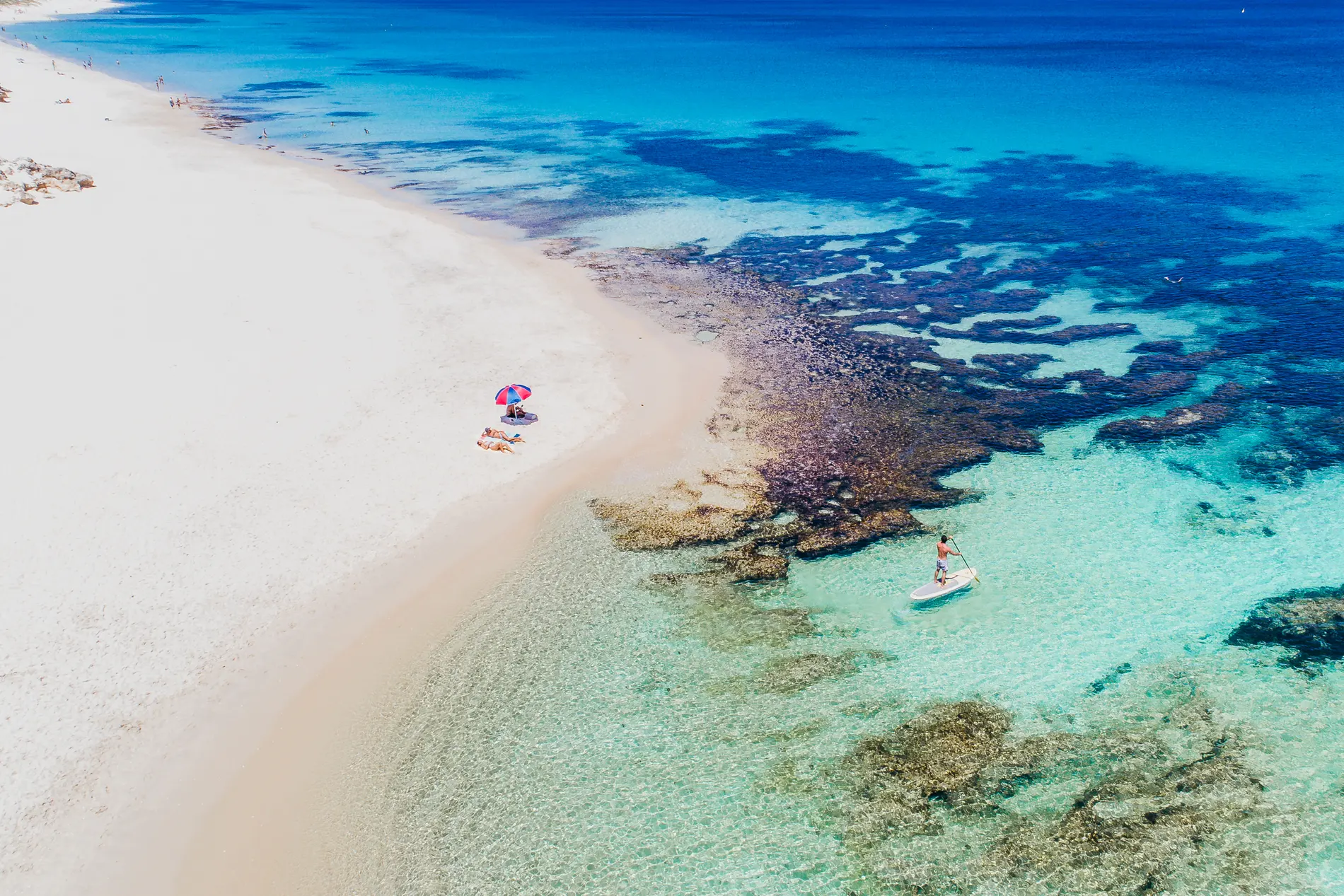 Aerial of a white sand beach meeting turquoise shallows and dark reef, with a paddleboarder offshore