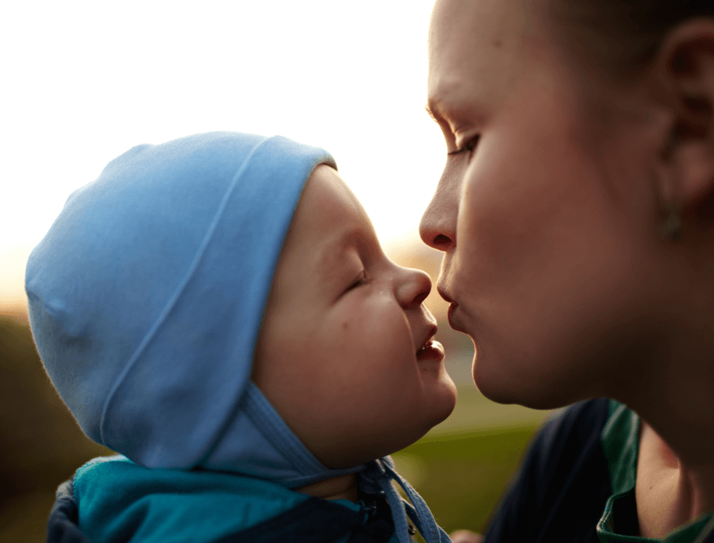 A woman and a baby share a playful kiss, both smiling, against a soft, natural background.