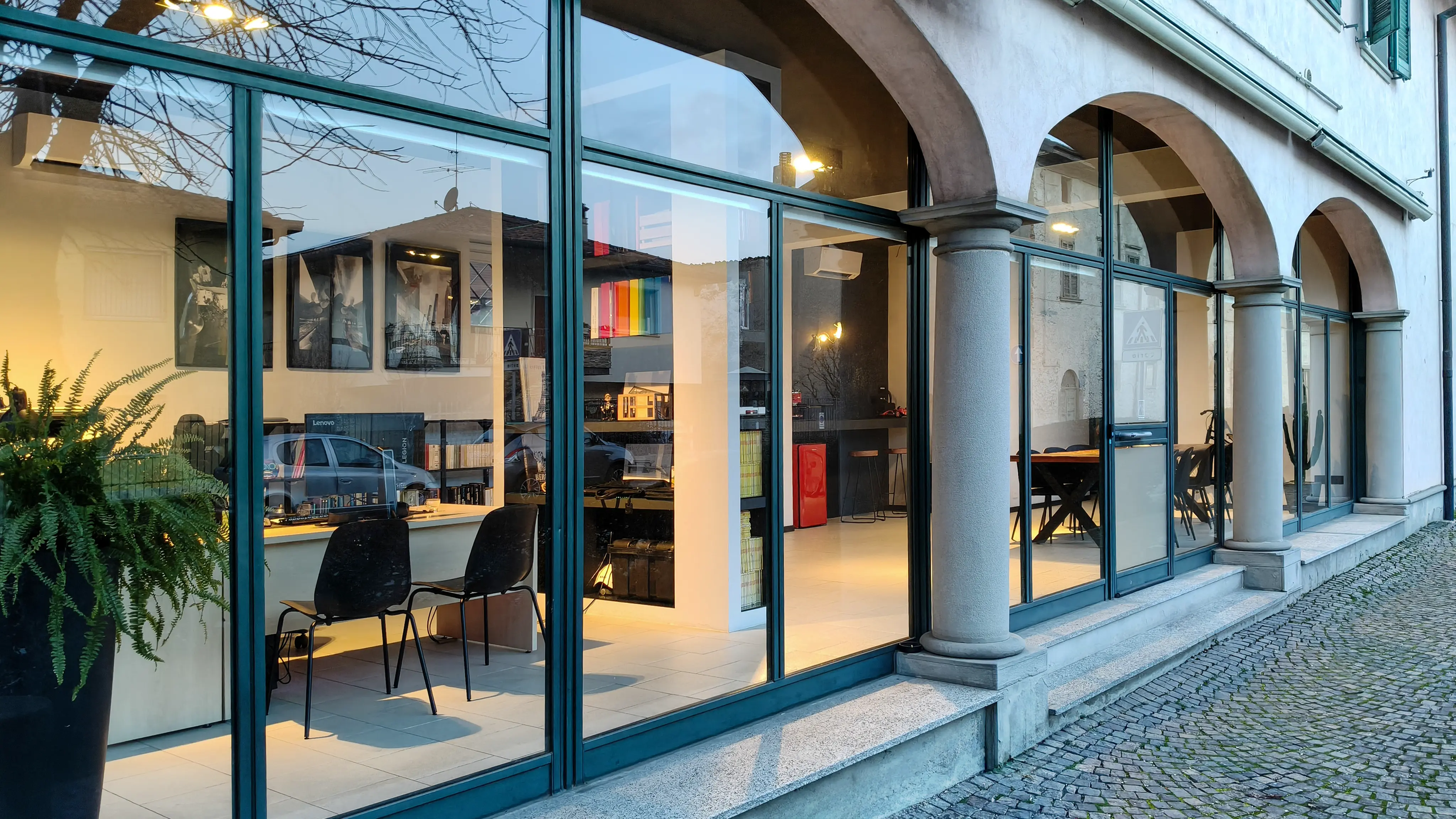 A spacious industrial-style interior with exposed brick walls, large windows, and natural light pouring in during sunset. Wooden desks with plants and workspace materials are arranged along the walls, with a view of greenery and an outdoor seating area.
