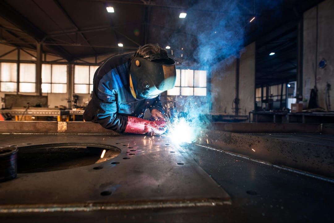 A worker is wearing a welding helmet and gloves. He welds a metal sheet, with bright sparks and smoke, inside a fabrication workshop.