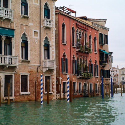 运河两岸矗立着带有拱形窗户和阳台的历史建筑，水中设有蓝白条纹的系船桩。