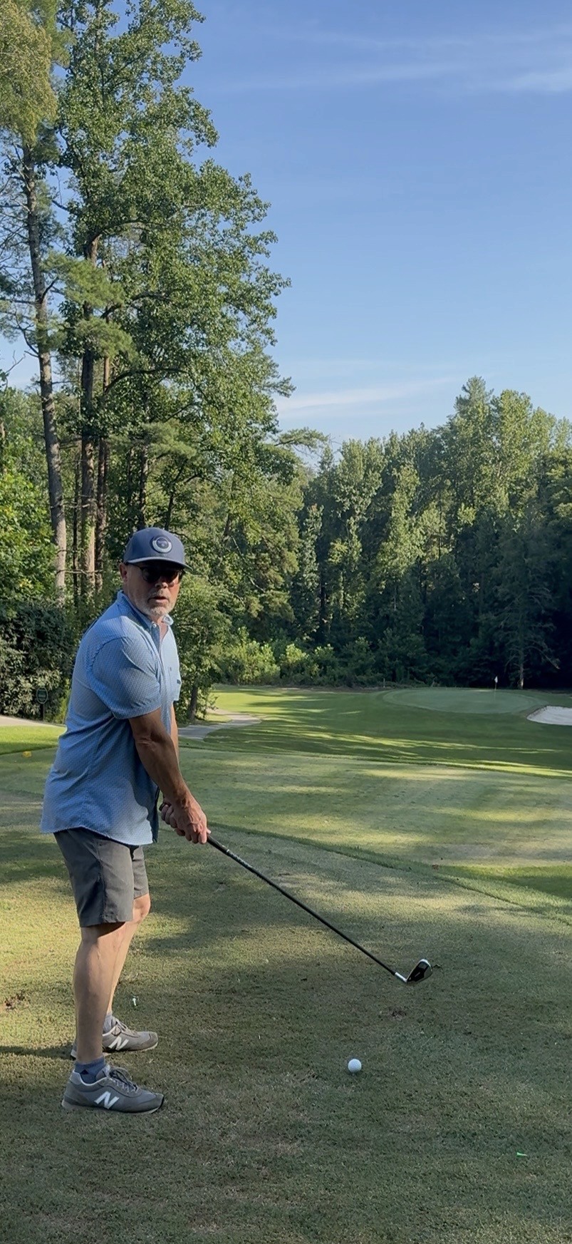 A golfer stands on the fairway preparing for an approach shot toward a green surrounded by tall trees beneath a bright blue sky.