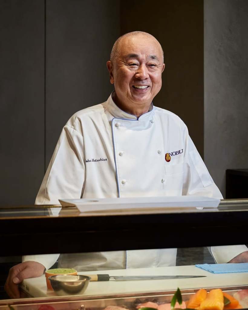 A smiling chef in a white uniform stands behind a sushi counter, showcasing his culinary expertise in a restaurant setting.