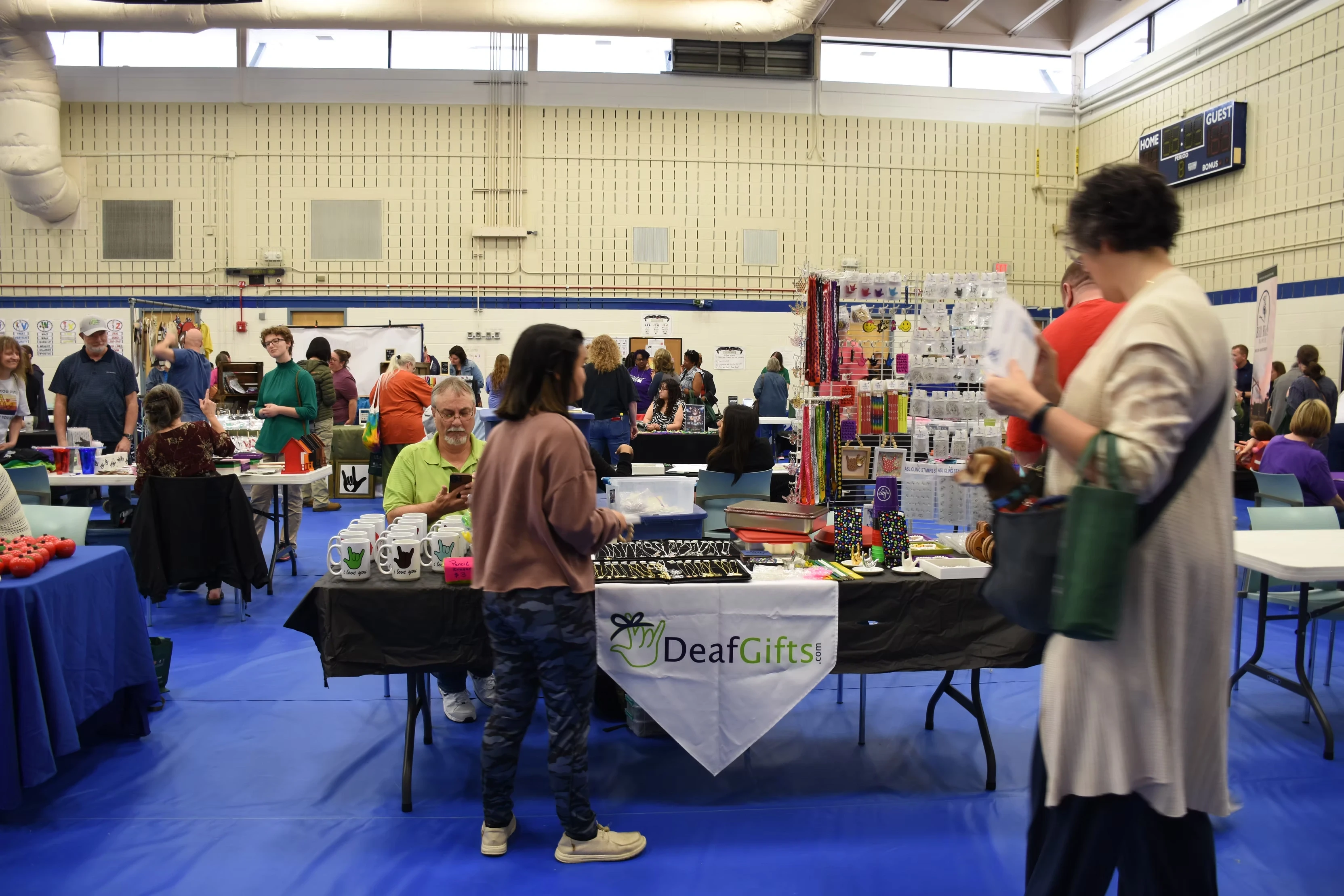 A craft fair in a gymnasium. Several vendors are at tables displaying their goods. A woman is looking at jewelry at the DeafGirls! booth.