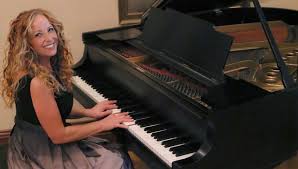 A smiling man with curly hair is sitting at a desk, looking at the camera, with a laptop and a lamp in the background.