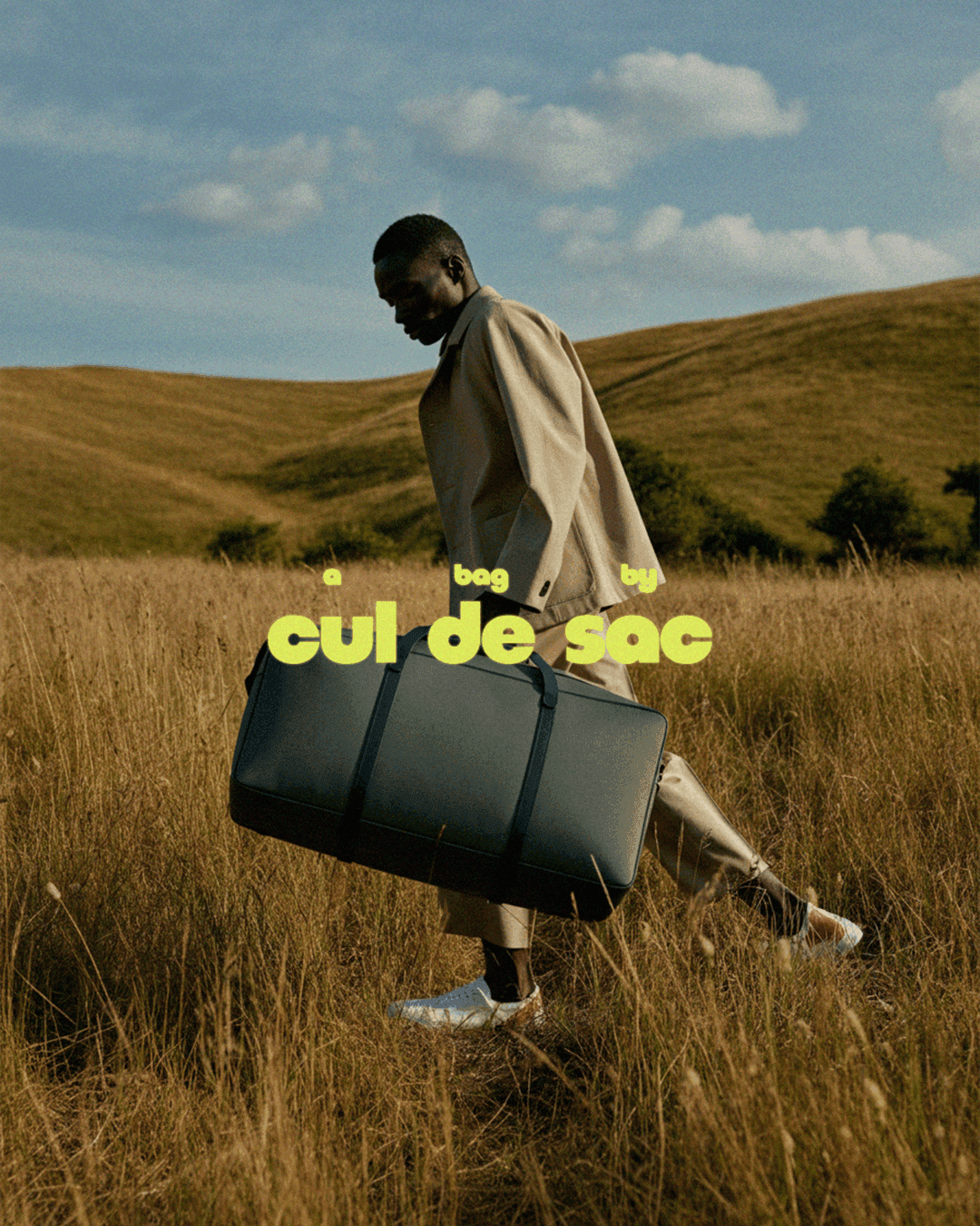 A person walking through open grassland carrying a large dark duffel bag, with the cul de sac wordmark overlaid in chartreuse
