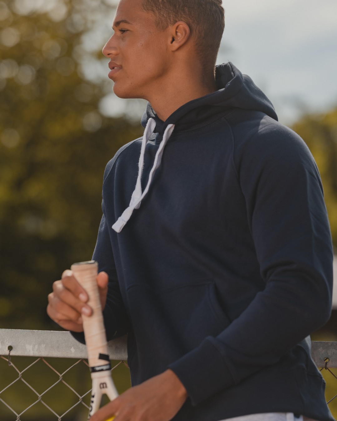 Person wearing a navy blue hoodie and holding a tennis racket outdoors in summer
