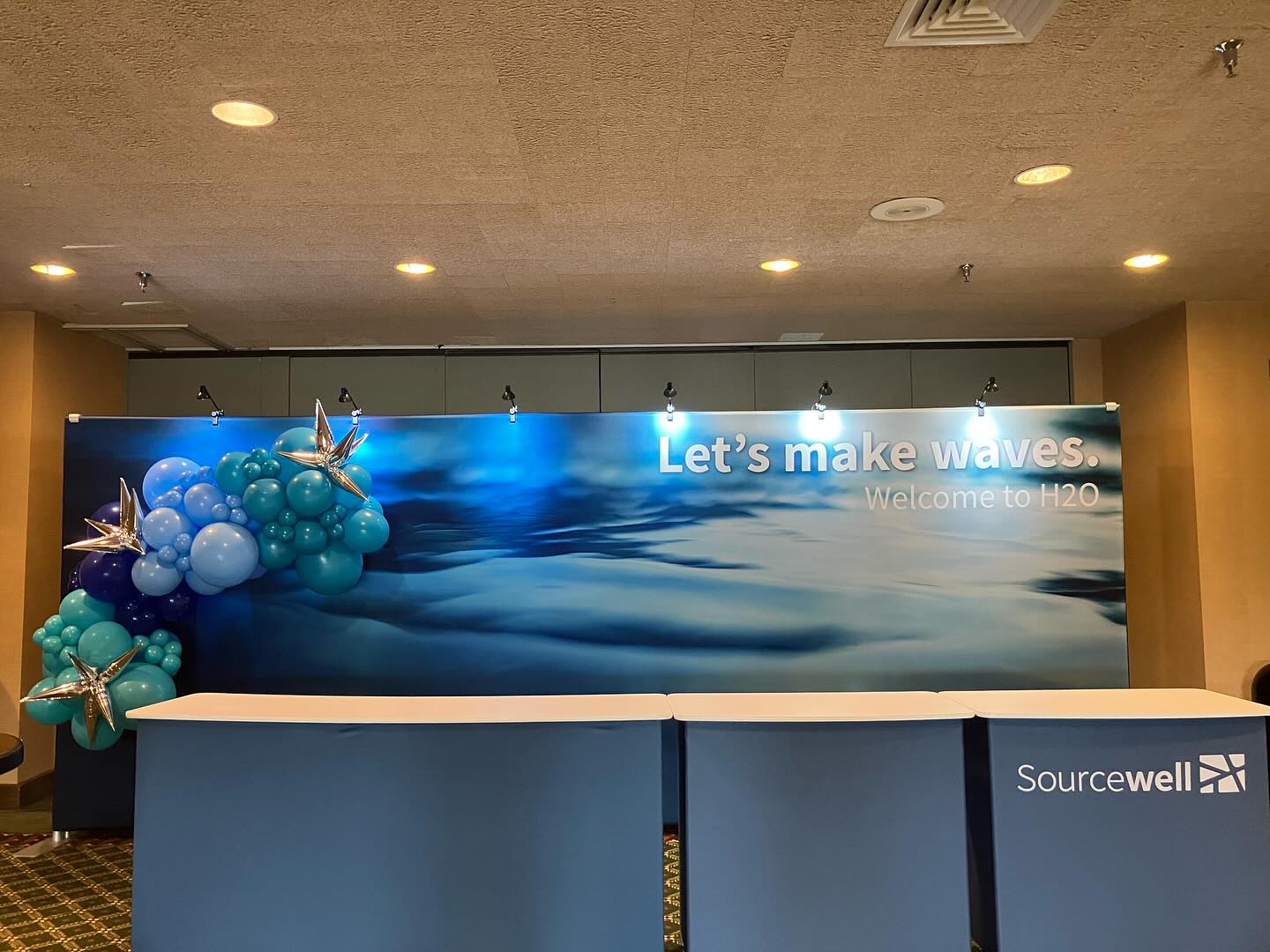 A conference booth display features a backdrop with a blue ocean wave design and the message "Let's make waves. Welcome to H2O," flanked by clusters of blue and teal balloons with silver star-shaped balloons, alongside two blue tables with "Sourcewell" branding.