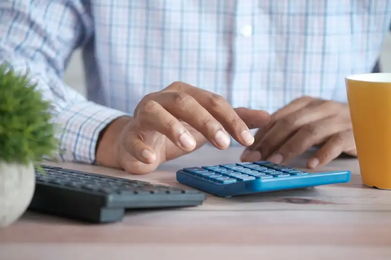 Gros plan sur les mains d'une personne en chemise à carreaux utilisant une calculatrice de bureau bleue sur une table en bois, à côté d'un clavier d'ordinateur et d'une tasse jaune.