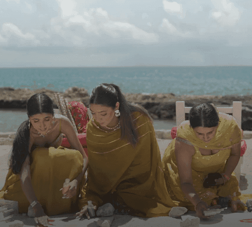 Three women in yellow dresses kneeling on a beach, arranging rocks into a pattern with large rocks, the coast, and crystal-clear water in the background.