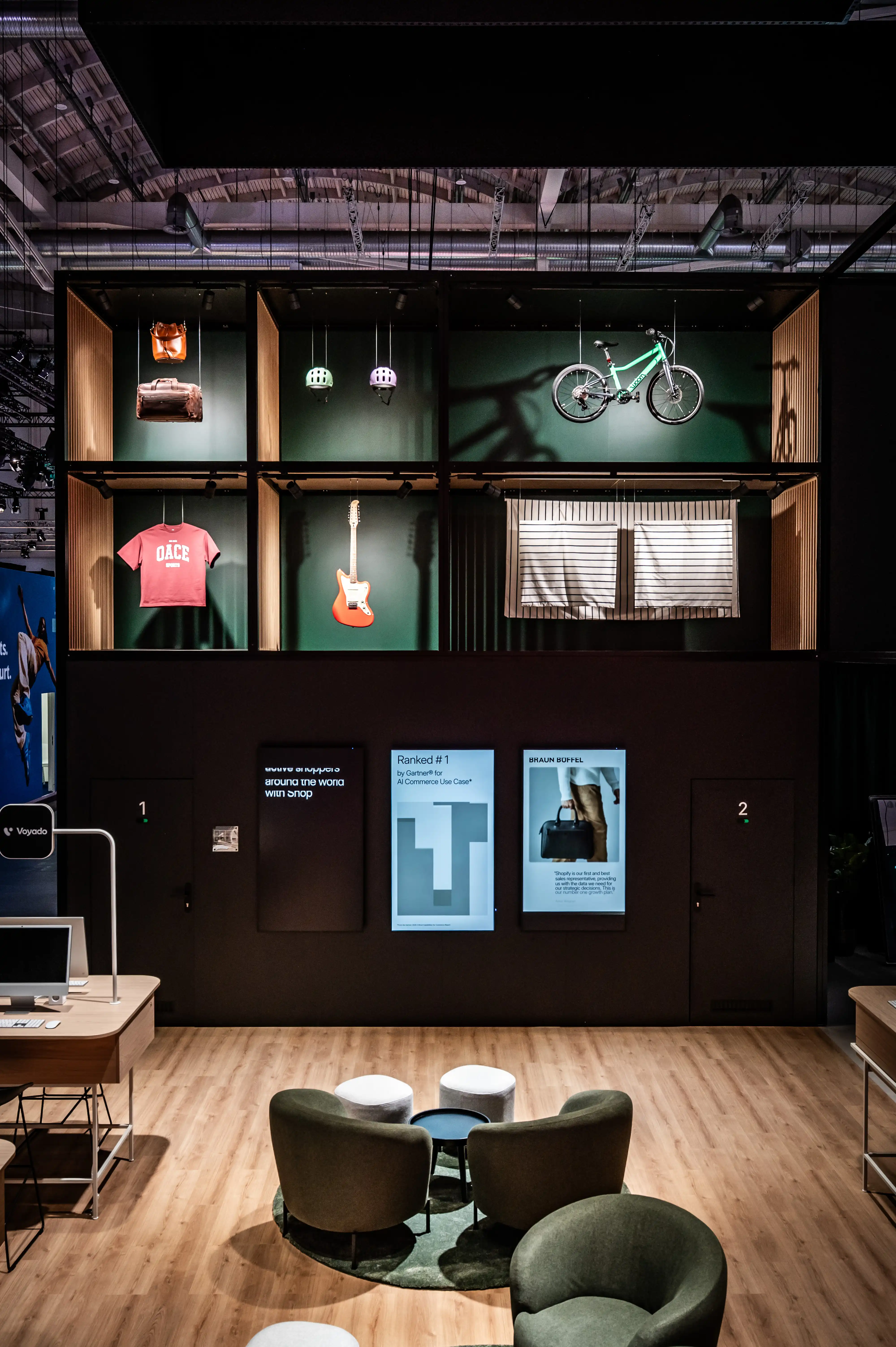 Tall green product shelf with guitar, bike, shirt and lamps above screens and lounge chairs.
