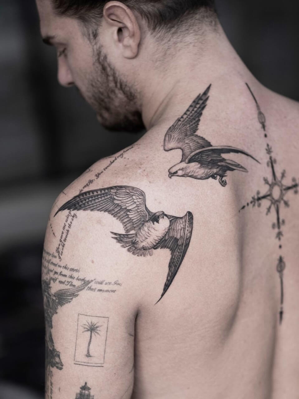 A man with various tattoos on his back and shoulder, featuring birds in flight, a palm tree, and cursive text. One of the prominent birds is detailed with outstretched wings.