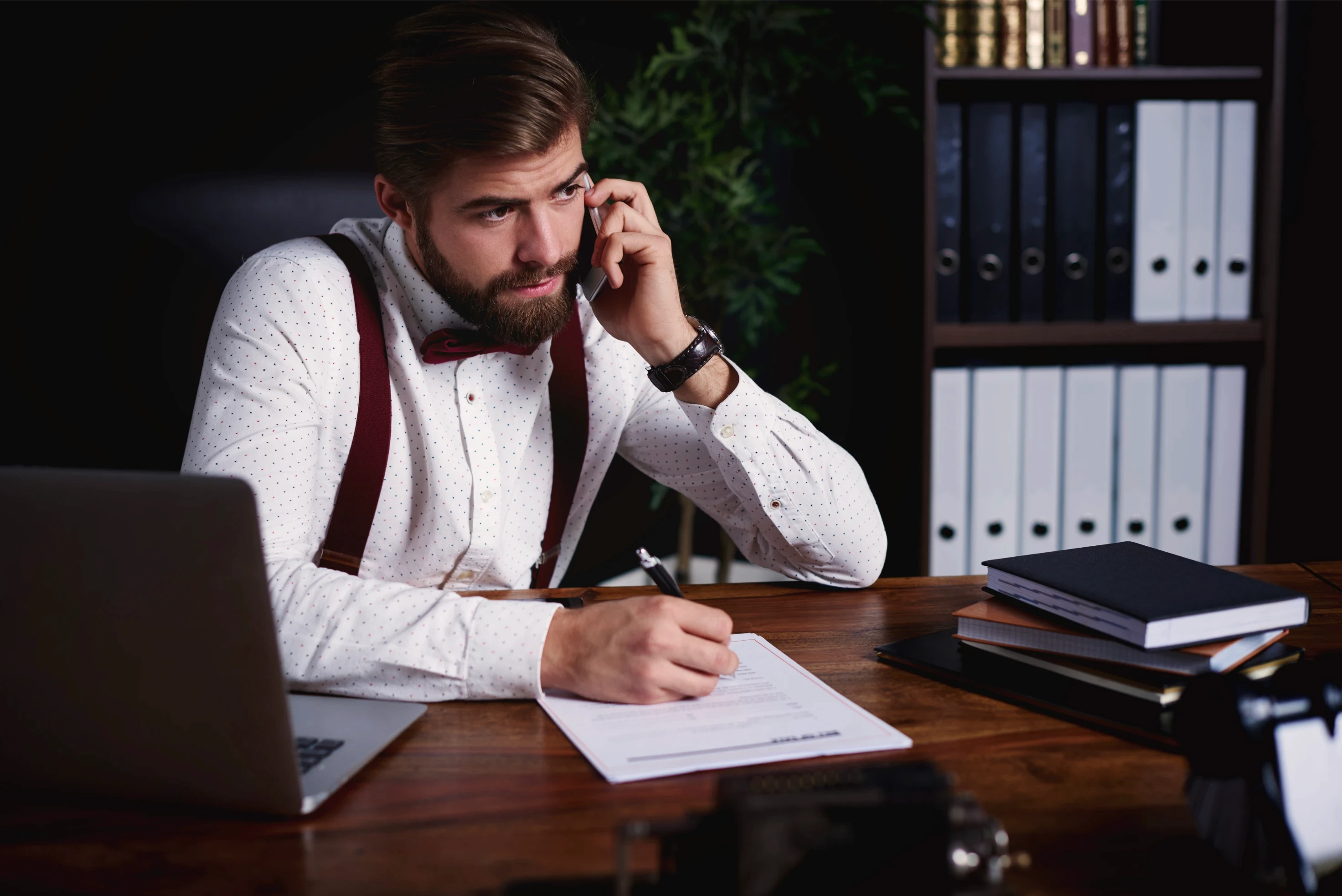 A man in a white shirt and burgundy suspenders works at a wooden desk, holding a phone to his ear and writing on a document, creating a focused and professional atmosphere.