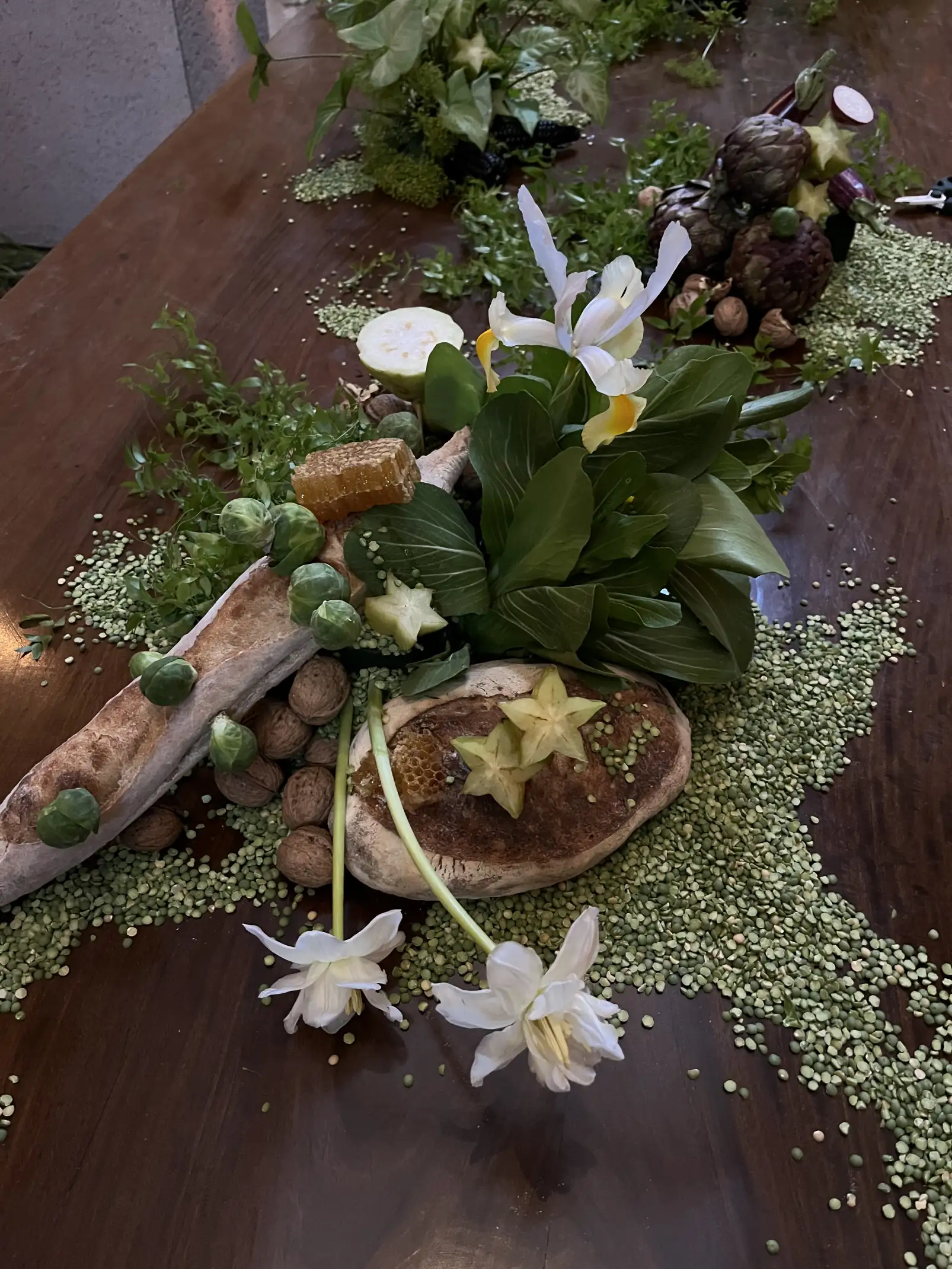 Composición naturalista de centro de mesa con flores blancas, pan rústico, nueces y guisantes verdes sobre superficie de madera.