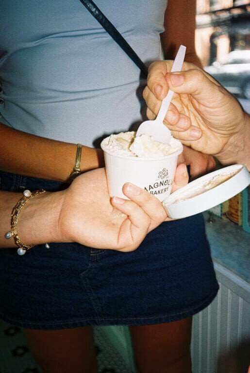 A flash photo of a woman holding a cup of magnolia bakery banana pudding as other people reach in frame to grab a spoonful