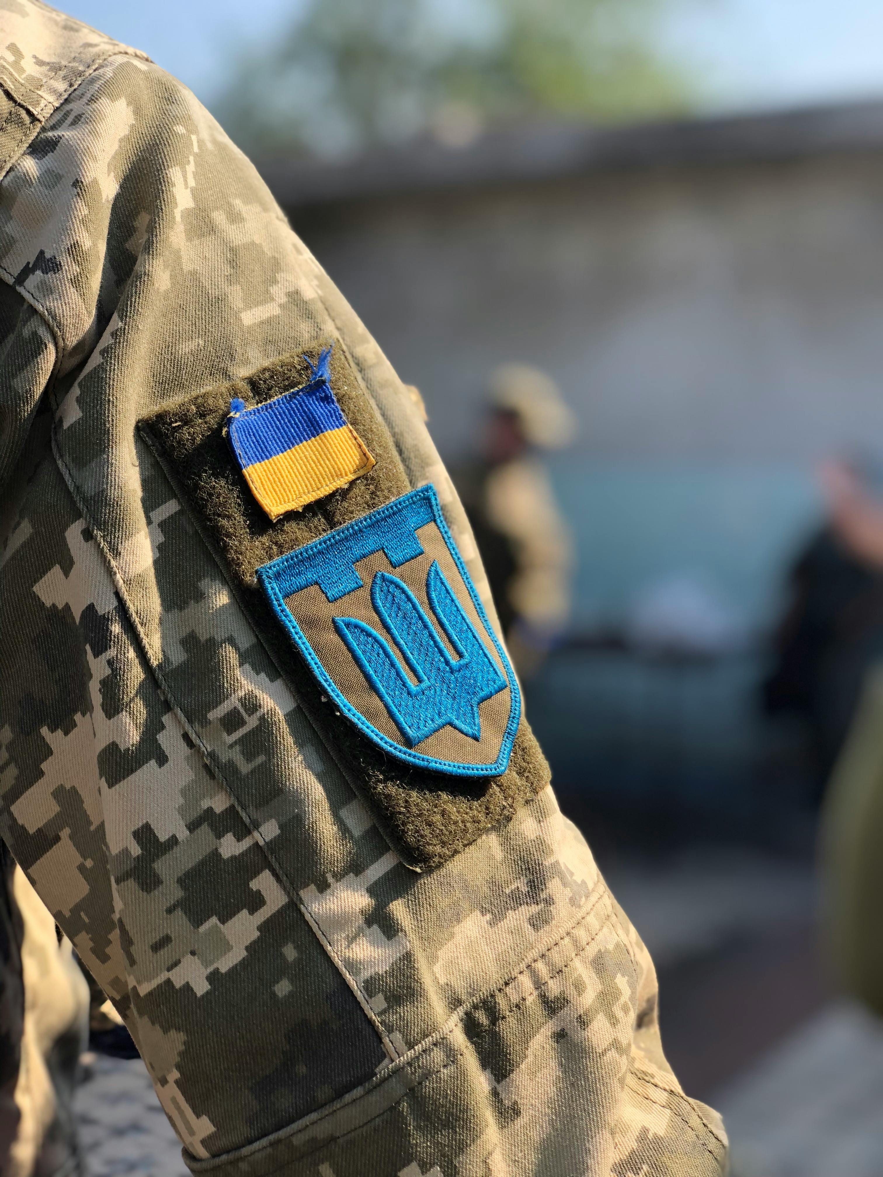 A soldier's arm bearing the Ukrainian flag and the Ukrainian coat of arms, the Tryzub. The flag symbolizes Ukraine's independence and national pride, while the Tryzub, a historical emblem, represents sovereignty and the country's cultural heritage.