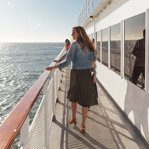 一位穿着牛仔夹克的女士漫步在船甲板上，右侧是碧海蓝天的美丽景色。
