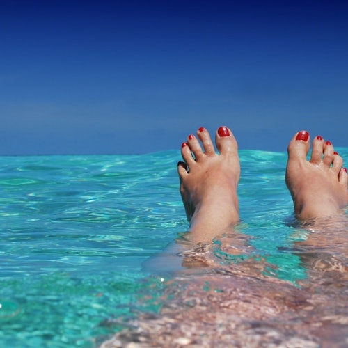 Feet with red toenails floating in clear turquoise water against a blue sky backdrop.
