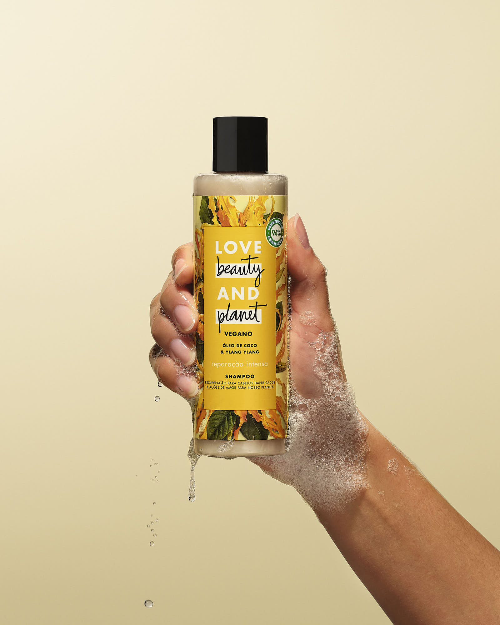 Hand holding a Love Beauty and Planet vegan shampoo bottle with coconut oil and ylang ylang, dripping water, against a beige background.