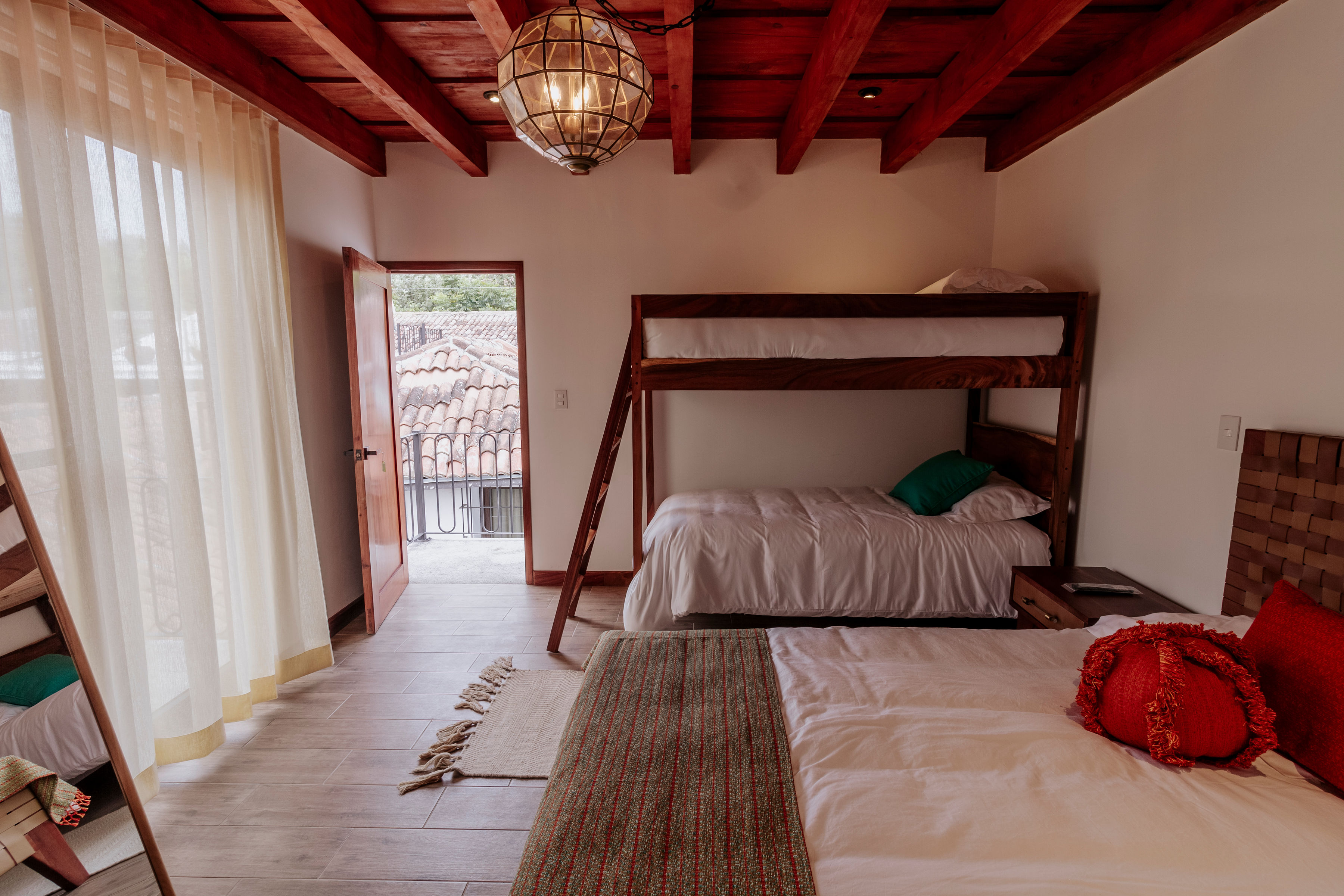 A cozy bedroom with wooden bunk beds and a double bed, featuring warm tones and rustic decor, illuminated by a sunlit glass door opening to a view of terracotta rooftops, enhancing the room's inviting atmosphere.